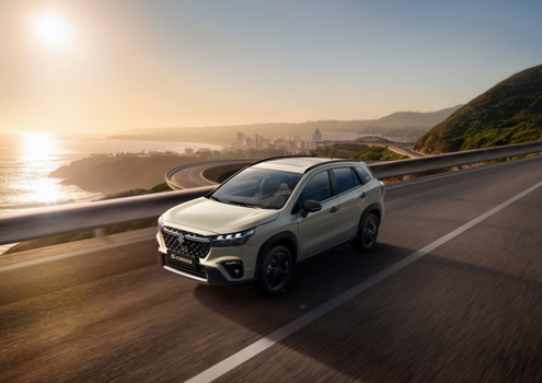 A silver S-Cross Kuro driving on a coastal road with a scenic sunset view.