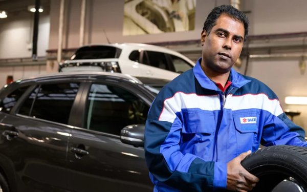 Mechanic in blue uniform holding a tyre in a garage, cars visible in the background.